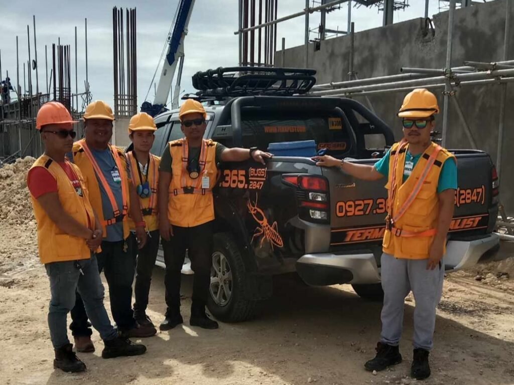 Updated Alt Text: Five exterminators from Technologist Pest Control, wearing orange safety vests and helmets, stand beside their branded black service vehicle labeled "TERMINATOR SPECIALIST," featuring a termite logo and contact number. They're on-site for a pre-construction termite proofing job, with visible scaffolding, steel rods, and a crane in the background.